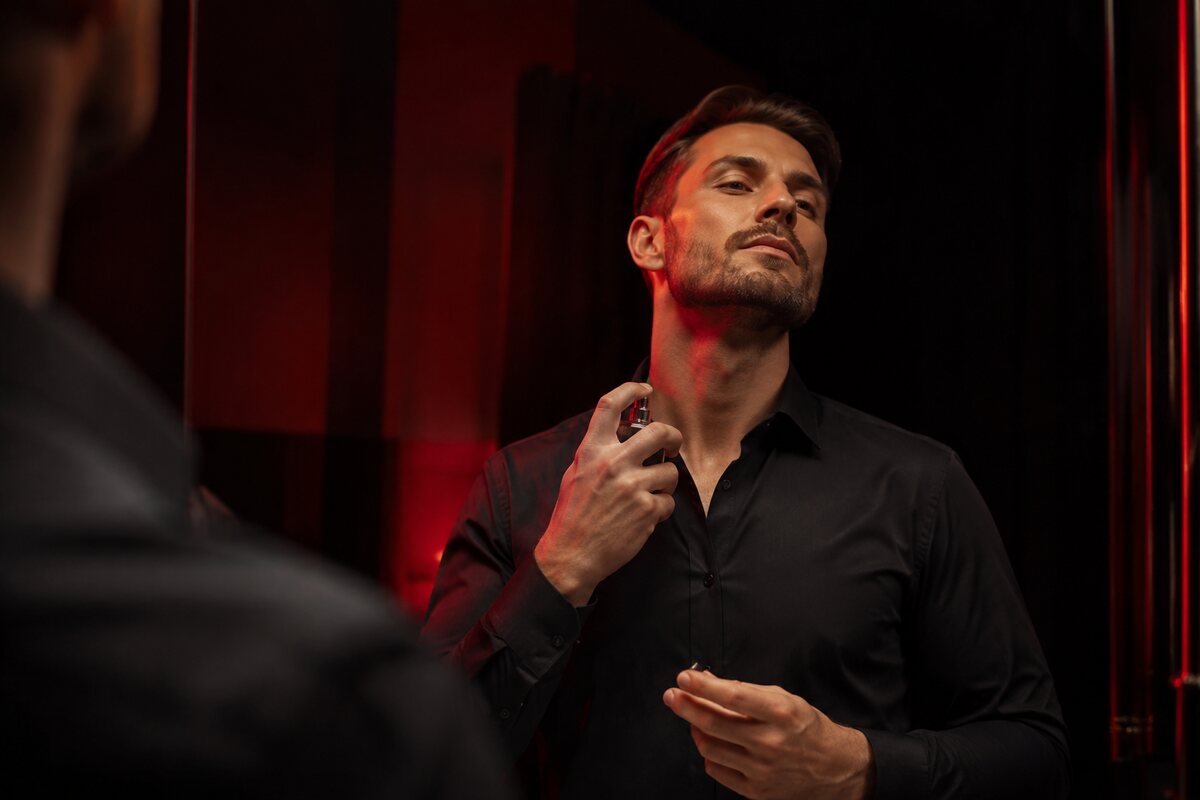 a well-groomed man applying fragrance after moisturizing clean skin in a dark red-lit dressing area